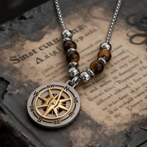 May include: A silver-tone compass pendant necklace with a gold-coloured compass rose design. The necklace features brown tiger eye beads and a chain. The compass pendant is approximately 3.8 cm in diameter.