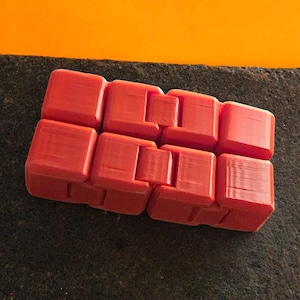Könnte beinhalten: Ein rotes Fidget-Spielzeug aus verbundenen Würfeln. Das Spielzeug besteht aus Kunststoff und hat eine strukturierte Oberfläche. Die Würfel sind rechteckig angeordnet und können manipuliert werden. Etwa 15 cm lang.