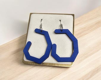 May include: A pair of geometric, blue wooden earrings. The earrings have a unique, angular design with a silver hook. They are displayed on a cream-colored surface inside a gold-toned box, set against a light wood background.