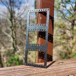 Op de afbeelding: Een zwarte metalen filmnegatiefhouder met drie rechthoekige openingen. De houder heeft een getextureerd oppervlak en staat op een houten oppervlak.