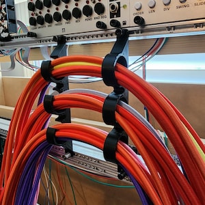 Puede incluir: Primer plano de una configuración de sintetizador modular con cables trenzados coloridos sujetos por soportes negros. Los cables son principalmente naranjas, rojos y morados, con acentos amarillos y otros colores. El sintetizador tiene perillas y etiquetas.