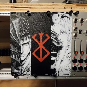 May include: A black and white image of a character from the anime series Berserk, with a red and white symbol in the background. The image is mounted on a silver panel with knobs and buttons.