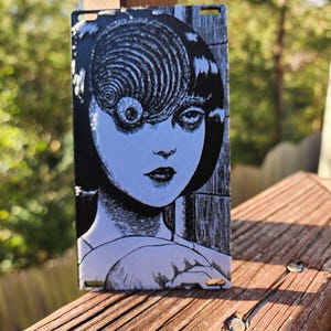 May include: A black and white metal plate with a graphic of a woman's face with a spiral eye. The woman's hair is black and her eyes are closed. The background is a textured surface.