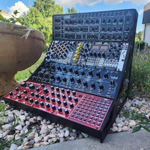 May include: A black and red synthesizer with a variety of knobs and buttons. The synthesizer is on a stand with other synthesizers stacked on top of it.