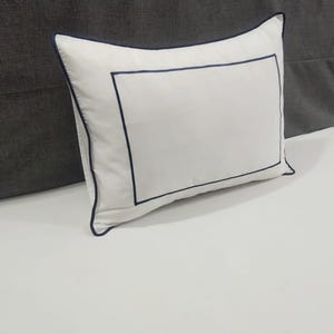Könnte beinhalten: Weißes rechteckiges Kissen mit marineblauem Rand und Zierleiste. Das Kissen liegt auf einer weißen Oberfläche. Das Design ist schlicht und elegant, passend für ein Schlafzimmer.