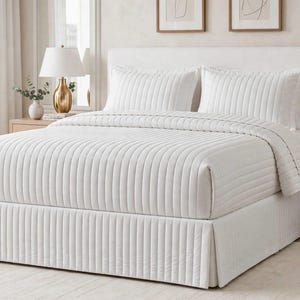 May include: A white bed set featuring a quilt and two matching pillow shams, all decorated with a vertical channel-stitched pattern. The bed is completed with a matching white pleated bed skirt that covers the base of the mattress.