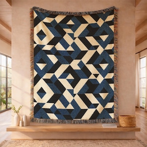 May include: A woven throw blanket featuring a repeating geometric pattern of interlocking cubes in shades of navy blue, black, and cream. The rectangular textile has a fringed edge along all four sides with multicolored threads visible.