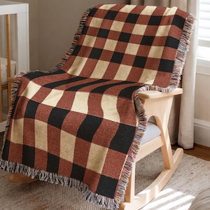 May include: A woven throw blanket with a large buffalo check pattern in black, tan, and rust red is draped over a wooden rocking chair. The blanket has a fringed edge and is placed on a light gray seat cushion.
