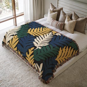 May include: A woven throw blanket with a repeating leaf pattern in yellow, green, navy blue, and cream colors. The blanket has a black background and a fringed edge. The blanket is displayed on a bed with pillows.