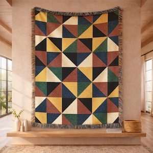 May include: A woven throw blanket featuring a geometric pattern of triangles in shades of mustard yellow, forest green, deep navy blue, terracotta red, black, and cream. The rectangular blanket has a fringed edge along all four sides.