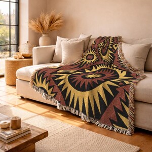 May include: A woven throw blanket featuring a repeating pattern of large, stylized sunburst shapes in shades of gold, deep red, and black. The blanket has a textured, fringed edge and is draped over a light-colored sofa with matching throw pillows.