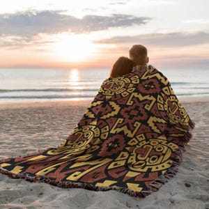 May include: A woven blanket with a geometric pattern in shades of brown, black, and yellow. The blanket is draped over two people on a sandy beach, with the ocean and sunset in the background. The blanket has a fringed edge.
