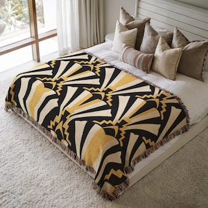May include: A woven throw blanket with a geometric fan pattern in black, cream, and gold. The blanket is fringed and placed on a bed with pillows. The bed is on a white carpet.