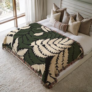May include: A woven throw blanket with a fern leaf design. The blanket features a cream-colored background with large green and black fern leaves. The edges are finished with a fringe.