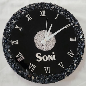 May include: A round black wall clock featuring silver Roman numerals, silver clock hands, and a central silver glitter circle. The name Soni is printed in silver below the centre. The outer edge is decorated with dark, textured crushed glass.