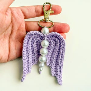 May include: A handmade lavender crochet angel wing keychain with a gold-colored clasp. The wings are adorned with a string of white pearl beads. The keychain is a decorative accessory.