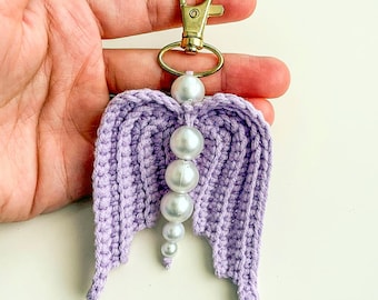 May include: A handmade lavender crochet angel wing keychain with a gold-colored clasp. The wings are adorned with a string of white pearl beads. The keychain is a decorative accessory.