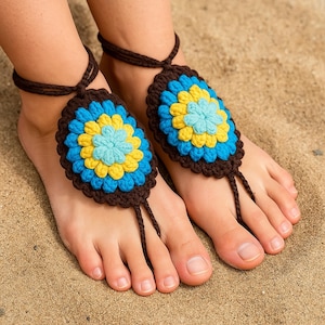 May include: Crocheted barefoot sandals featuring a floral design. The sandals have a brown border, with blue, yellow, and light blue flower centers. The ankle straps and toe loops are brown. The sandals are displayed on a sandy surface.