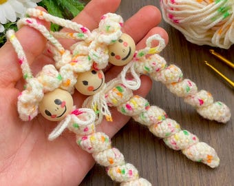 May include: Handmade crochet dolls with wooden bead faces. The dolls have a cream-colored body with colorful flecks and a smiling face. The yarn is cream-colored with flecks of pink, orange, and green. The dolls are a whimsical craft item.