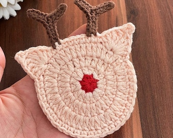 May include: A handmade, crocheted reindeer coaster in a light beige color. The coaster features brown antlers and a red nose. The design is circular with a reindeer face shape. The coaster is likely for decorative or functional use.