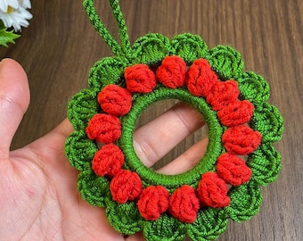 May include: A handmade crochet wreath with a green base and red berry accents. The wreath has a loop for hanging and is held in a hand. The wreath is a festive decoration.