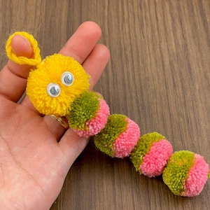 Pode incluir: Uma lagarta feita à mão com pompons amarelos, verdes e rosa. A cabeça da lagarta é amarela com olhos móveis. O corpo é uma série de pompons em cores alternadas verde e rosa. Um artigo de artesanato.
