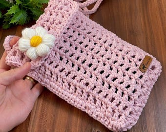 May include: A handmade, light pink crochet bag with a daisy embellishment. The bag features an open weave design and a small strap. A small leather tag with the word "Handmade" is attached to the bag.