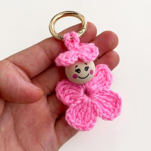 May include: A handmade pink crochet flower keyring with a wooden bead face and gold-coloured ring. The flower has a smiling face with rosy cheeks and a matching pink hat. The keyring is a charming accessory.