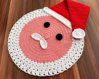 May include: A handmade, crocheted Santa Claus face mat. The mat features a pink face with black eyes, white mustache, and a red and white Santa hat. The mat is round with a white border.