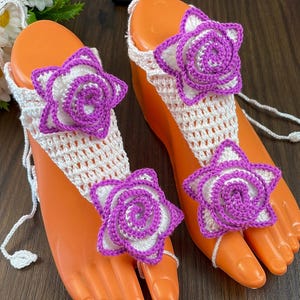 Puede incluir: Sandalias descalzas de ganchillo blanco con detalles florales morados y blancos. Las sandalias presentan un diseño de correa en T y están adornadas con adornos florales tridimensionales. Las sandalias se exhiben en formas de pies naranjas.