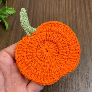 May include: A handmade, orange crochet pumpkin coaster with a green stem. The coaster is round and has a textured surface. The stem is a light green colour. The coaster is approximately 10 cm in diameter.