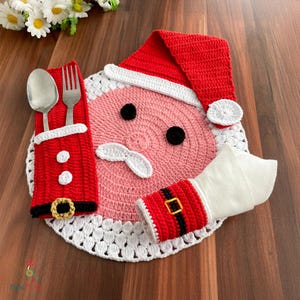 May include: A festive Christmas table setting with a crocheted Santa Claus design. The set includes a red and white Santa hat, a pink face with black eyes, and a white mustache. A red utensil holder and napkin holder complete the festive look.
