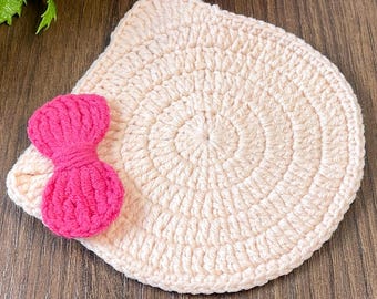 May include: A circular, cream-colored crocheted coaster or mat featuring a textured, concentric stitch pattern. Attached to the left side is a small, bright pink crocheted bow with a ribbed, vertical texture, adding a pop of color.