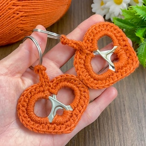 Puede incluir: Dos llaveros de ganchillo naranja con forma de pretzel con anillos de metal plateado. Los llaveros están hechos con hilo naranja y presentan una anilla plateada en el centro. En el fondo se ve una madeja de hilo naranja.