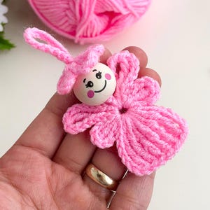 May include: A handmade pink crochet angel ornament with a wooden bead face featuring painted eyes and a smile. The angel has a flower-shaped body and a loop for hanging. A ball of pink yarn is in the background.