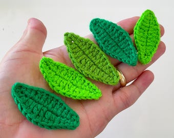 May include: Six handmade crochet leaves in varying shades of green. The leaves are arranged in a fan shape, showcasing the different textures and colors of the yarn. The leaves are small, and appear to be decorative.