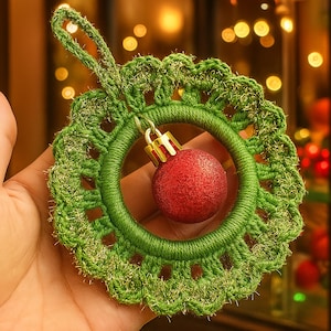 Può includere: Un ornamento natalizio fatto a mano con una ghirlanda verde all'uncinetto con una palla rossa glitterata al centro. L'ornamento ha un anello per appenderlo. La ghirlanda è decorata con filo d'oro.