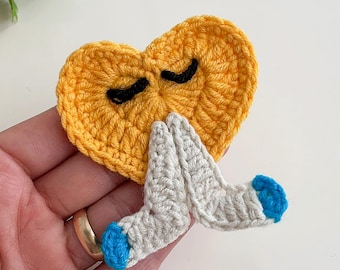 May include: A crocheted heart-shaped patch in yellow with black eyelashes and praying hands. The hands are gray with blue tips. The patch is handmade and likely used for clothing or accessories.
