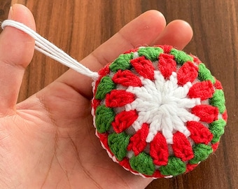May include: A crocheted Christmas ornament in a ball shape. The ornament features a pattern of red, green, and white yarn. A white string is attached for hanging. The ornament is held in a hand against a wooden background.