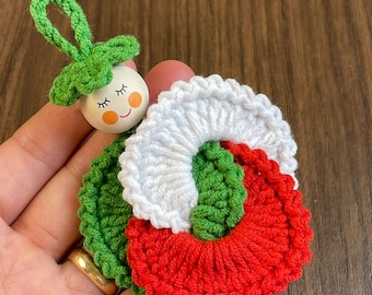 May include: A handmade Christmas ornament featuring a smiling wooden face with closed eyes, a green leaf-like top, and a crocheted wreath in red, white, and green. The ornament is held in a hand.