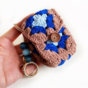 May include: A crocheted square pouch in a granny square pattern. The pouch features a color scheme of brown, blue, and light blue. A wooden button secures the flap. A gold-colored clasp and beaded charm are attached.