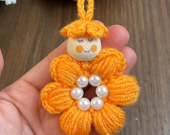 May include: A handmade orange yarn flower with a wooden doll face center. The doll has painted eyes, rosy cheeks, and a smiling mouth. The flower petals are made of looped yarn, and the center has white pearl beads. A loop of yarn is at the top.