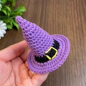 May include: A crocheted purple witch hat with a black band and a yellow buckle. The hat features a pointed crown and a wide brim. Ideal for Halloween or fancy dress events.
