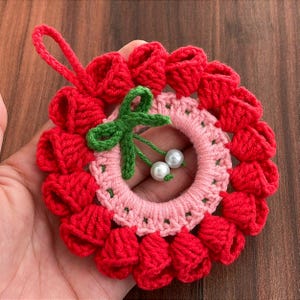 Puede incluir: Una corona de ganchillo hecha a mano con flores rojas en forma de campana alrededor de un centro rosa. Un lazo verde y dos perlas blancas están en el centro. La corona está sostenida por una mano.