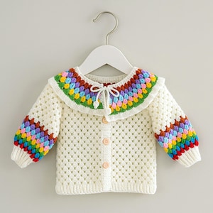 May include: A cream-colored, crocheted baby cardigan with a colorful rainbow collar and sleeves. The collar and sleeves feature a vibrant pattern of red, orange, yellow, green, blue, and purple. The sweater has wooden buttons and a delicate tie closure.