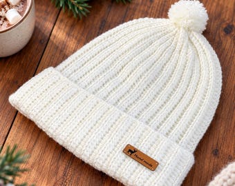 May include: A cream-colored ribbed knit beanie with a matching pom-pom on top and a folded brim. A small rectangular brown leather tag is attached to the front of the brim, featuring a black deer silhouette and the text "Hand made".