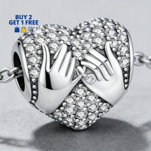 Puede incluir: Un charm de plata en forma de corazón con un diseño de dos manos sosteniendo un anillo con una gema transparente. El charm está incrustado con pequeñas gemas transparentes. El texto "BUY 2 GET 1 FREE" está en la esquina superior izquierda.