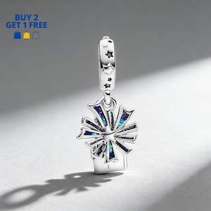 Puede incluir: Un charm de plata con un diseño de estrella y explosión de estrellas. El charm presenta una base rectangular con un diseño de explosión de estrellas en tonos azules y turquesas. La parte superior del charm tiene un lazo con recortes de estrellas.