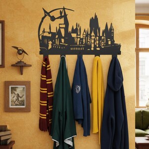 Könnte beinhalten: Ein schwarzer Wand-Kleiderhaken aus Metall mit einer Silhouette eines Schlosses und eines Uhrturms. Er hält einen gestreiften Schal, eine grüne Jacke, einen blauen Pullover und einen gelben Schal. Die Wand ist warmgelb gestrichen.