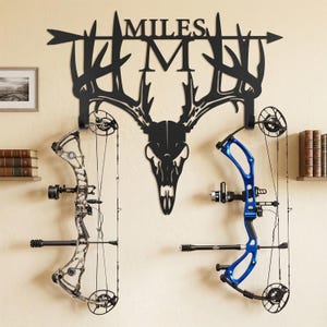 May include: Black metal wall art featuring a deer skull and the name "MILES" with an arrow. Two compound bows, one silver and one blue, are mounted below. The wall art is a decorative piece for archery enthusiasts.